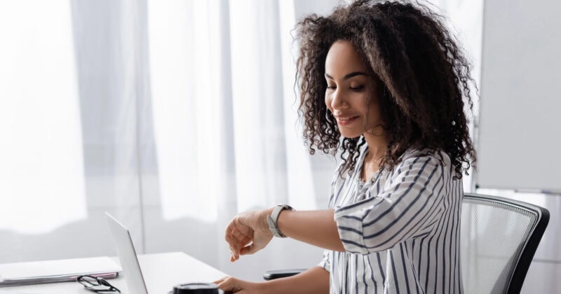 Woman sitting at a desk with a laptop, looking at her watch. A coffee cup, notebook, and calculator are on the desk.