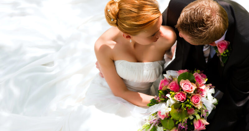 Bride in a white dress holding a bouquet of pink and white flowers sits closely with the groom in a black suit, both looking at each other—a picture-perfect moment for wedding websites.
