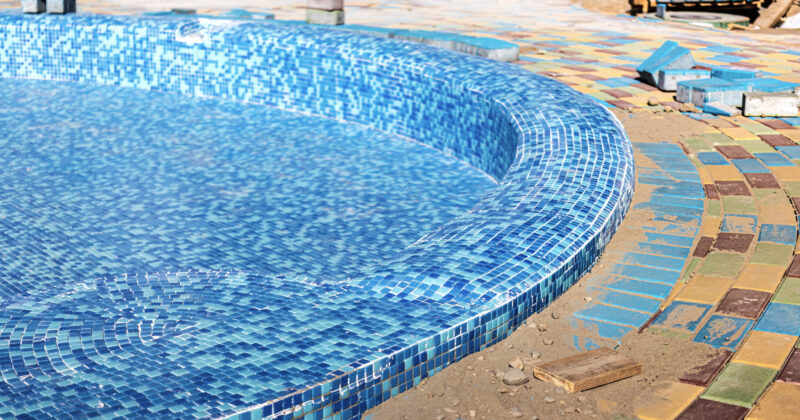 A close-up of a tiled swimming pool under construction by a skilled pool builder, with blue mosaic tiles inside the pool and multicolored tiles around the edge. Loose tiles and sand are visible nearby.