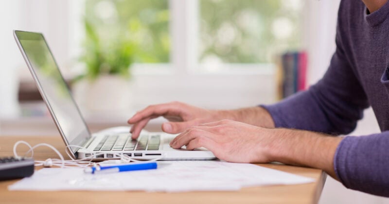 A person typing on a laptop at a desk with papers, a pen, and earphones scattered on the surface.