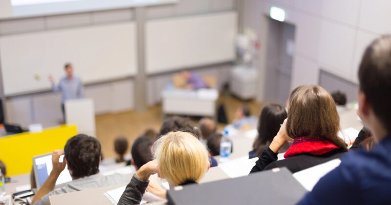 A group of students is seated in a lecture hall, facing a lecturer at the front near a podium and whiteboard—a classic scene that often features on the Best School Websites.