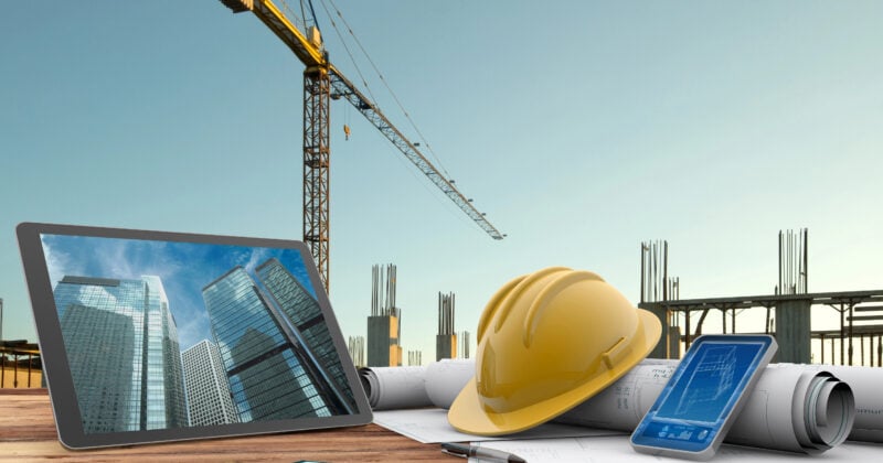 Tablet, hard hat, and architectural plans on a table at a construction site, with a crane and building framework in the background.