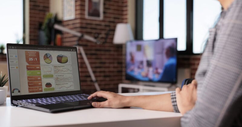 Person sitting at a desk using a laptop displaying charts and graphs; a television is on in the background in a modern room.
