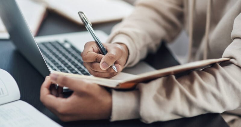 A person in a beige sweater jots down notes in a notebook while working on their laptop, seamlessly managing appointment scheduling with WordPress plugins.