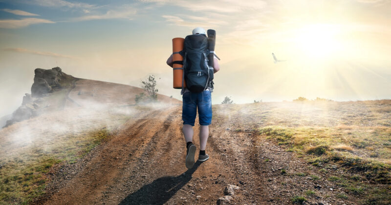 A person with a large backpack and camping gear walks alone on a dirt trail toward a rocky hill in bright, misty sunlight, symbolizing the buyer’s journey from search to sale.