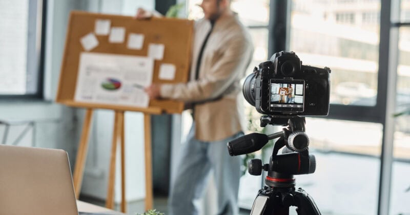 A man stands by a corkboard giving a presentation while being recorded on a video camera; the camera’s screen shows his image.