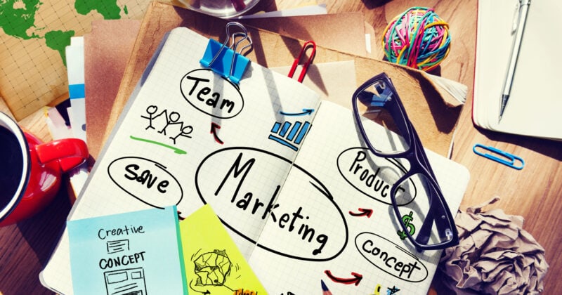 A desk showcasing an innovative marketing concept filled with creativity. The notebook is surrounded by essentials like glasses, colored pencils, sticky notes, a world map, a cup, and crumpled paper—perfect for startups crafting dynamic marketing strategies.