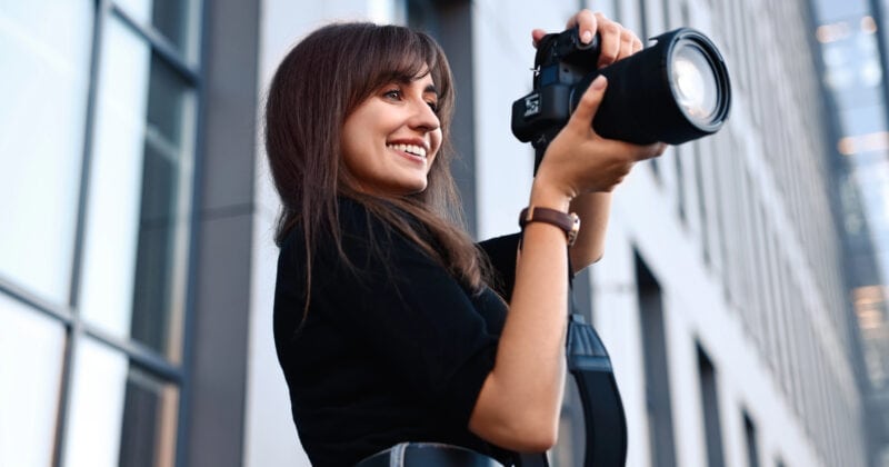 Woman holding a camera with a large lens, smiling while taking a photo in an urban setting with modern buildings in the background.