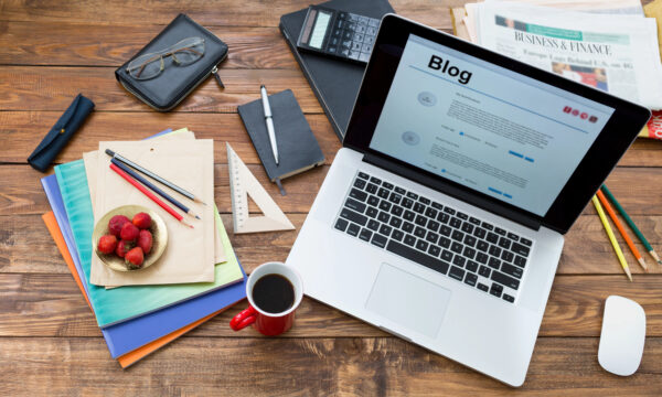A laptop displaying a blog, strawberries in a bowl, coffee in a red mug, pens, folders, a notebook, and a business newspaper are on a wooden desk.