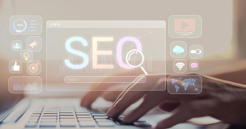 Hands typing on a laptop keyboard with a transparent overlay displaying the word "SEO" and various digital marketing icons.