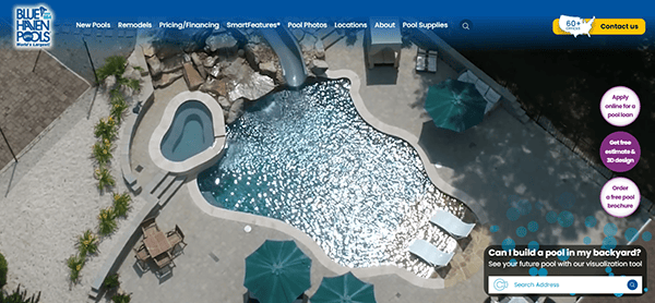 Aerial view of a landscaped backyard with a freeform swimming pool, spa, slide, lounge chairs, and umbrellas. Surrounding greenery and Blue Haven Pools’ expertly crafted pool website design are visible in the navigation at the top.