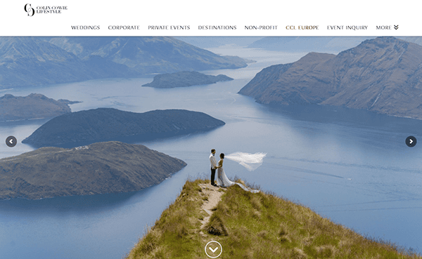 A bride and groom stand on a grassy cliff overlooking a lake and mountains, with the bride’s veil blowing in the wind—a dreamy moment often featured on the best wedding planner websites.