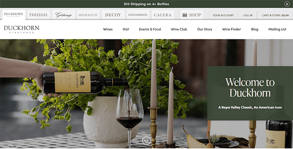 A hand pours red wine from a Duckhorn Vineyards bottle into a glass beside a potted plant, candles, and a "Welcome to Duckhorn" sign on one of the best winery websites.