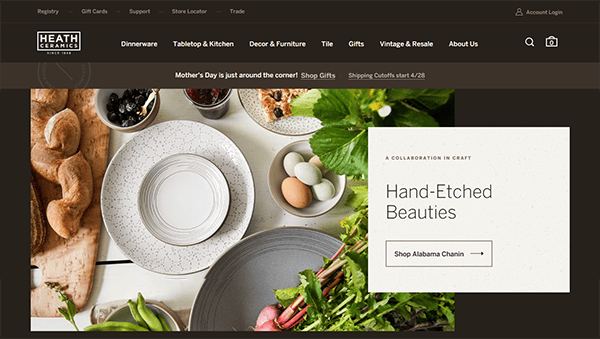 A table set with white speckled dishes, cutlery, bread, vegetables, and drinks, alongside a website banner promoting hand-etched ceramics, unique tile designs, and Mother's Day gifts.