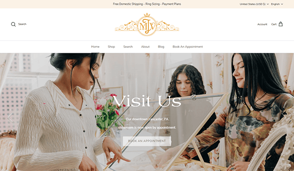 Two women work on jewelry displays in a boutique specializing in the best vintage clothing. Text overlay reads “Visit Us – Our downtown Lancaster, PA showroom is now open by appointment. Book An Appointment.”.