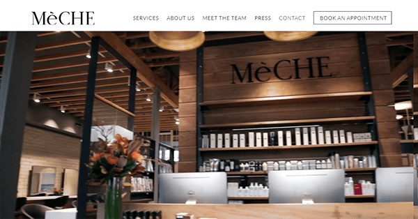 Modern salon interior with wooden accents, two computer monitors at the front desk—perfect for showcasing top salon website design—shelves of hair products, and a large "MeCHE" sign on the back wall.