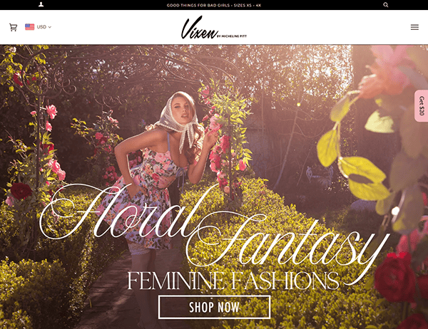 A woman in a floral dress poses in a garden surrounded by pink flowers. Text on the image reads "Floral Fantasy Feminine Fashions. Shop the best vintage clothing now.