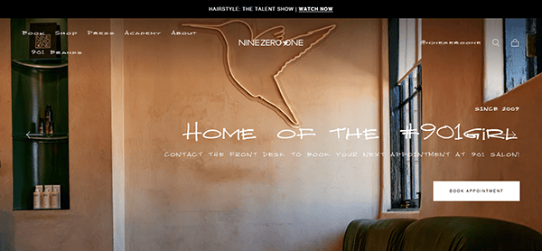 Interior of a salon with beige walls, a green couch, a built-in shelf, and a large hummingbird logo on the wall—showcasing the inviting atmosphere featured on the best salon websites. Book your appointment at 901 Salon today!.
