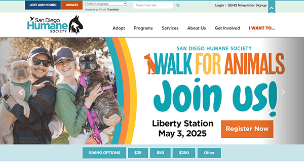 Two people holding dogs and smiling stand next to a sign promoting the San Diego Humane Society's "Walk for Animals" event at Liberty Station on May 3, 2025. Discover best pet adoption opportunities and register today!.