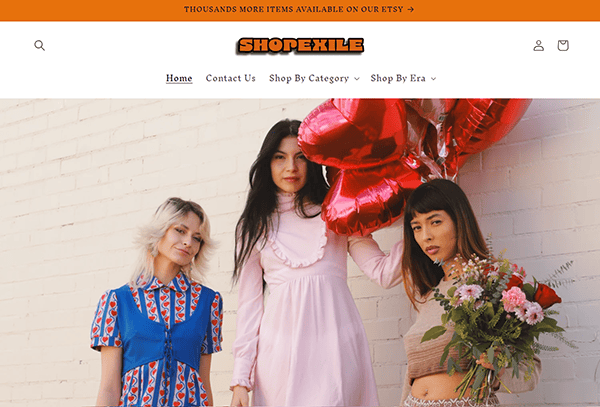 Three women pose in vintage clothing against a white brick wall; one holds heart-shaped balloons, another carries a bouquet of flowers, capturing the timeless flair found on the best vintage clothing websites.