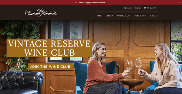 Two women sit on blue chairs clinking wine glasses in a wood-paneled room with a sign promoting the Vintage Reserve Wine Club at Chateau Ste. Michelle, an example often featured on the best winery websites.