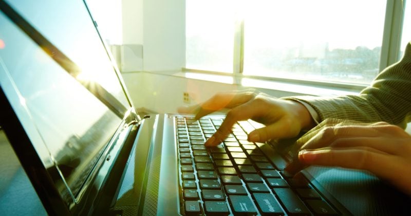 A person types on a laptop keyboard near a bright window with sunlight streaming in, exploring secure WordPress plugins for managing digital downloads.