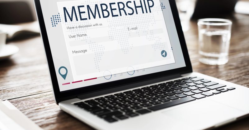 A laptop on a wooden table displays a WordPress membership registration form, featuring fields for username, email, and message. A glass of water and notebook sit beside the laptop, suggesting efficient use of membership plugins for a subscription model.