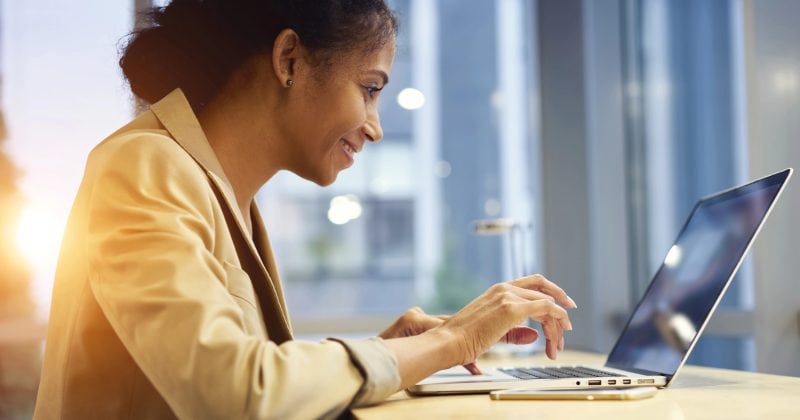 A woman sits at a desk, smiling as she types on a laptop in a bright, modern indoor setting—perhaps working on responsive menus with a menu plugin for WordPress.