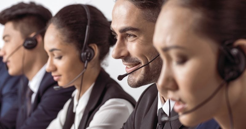 Four call center employees wearing headsets sit in a row, focused on their work, with one man smiling at his computer screen.