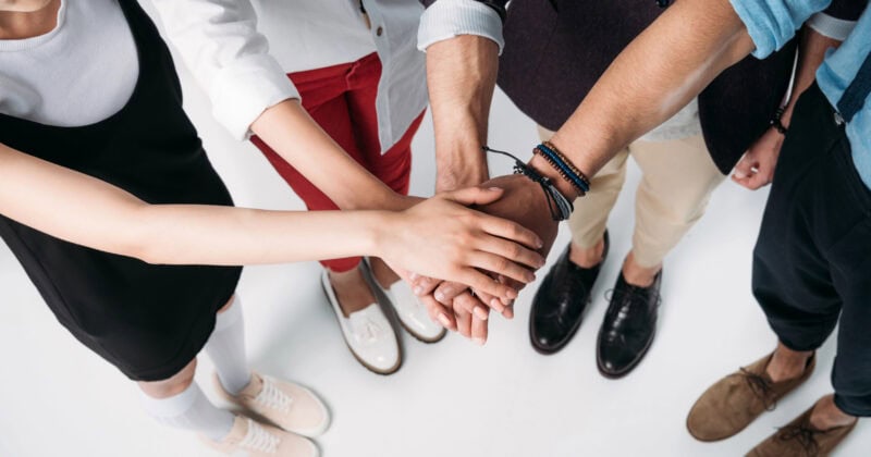 Four people standing in a circle stack their hands together in a gesture of teamwork, photographed from above on a white background.