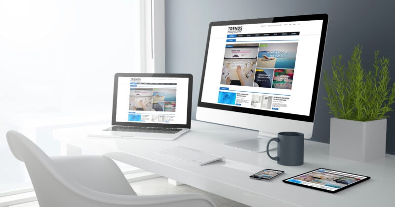 A modern white desk with a computer, laptop, tablet, and smartphone displaying the same website, next to a plant, a mug, and a notepad in a bright office space.