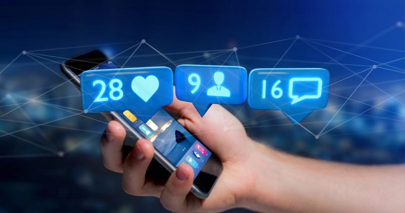 A hand holds a smartphone displaying social media notification icons—28 likes, 9 followers, and 16 messages—against a digital network background, illustrating rising user engagement through Facebook Feed Plugins on WordPress.