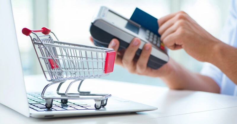A small shopping cart sits on a laptop, while a person holds a credit card near a card reader in the background.