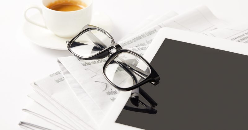 A pair of eyeglasses rests on a tablet placed on top of newspapers, with a cup of espresso in the background.