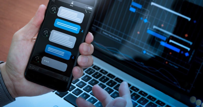 A person holds a smartphone displaying a chat app, with a laptop showing a project management or data visualization chart in the background.