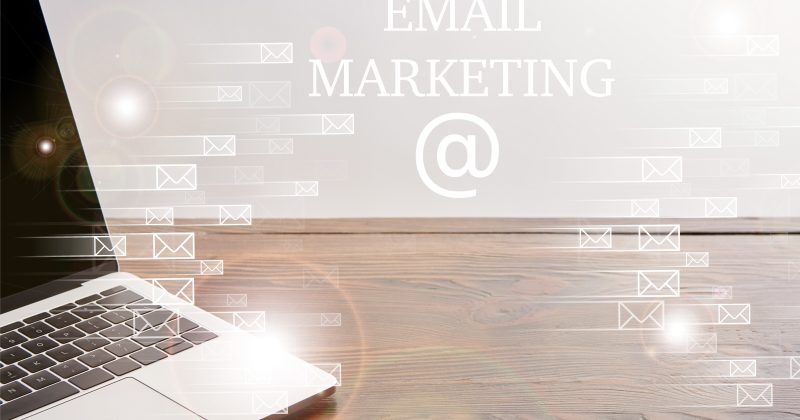 A laptop on a wooden desk with digital envelope icons and the words "Email Marketing" displayed in the background.