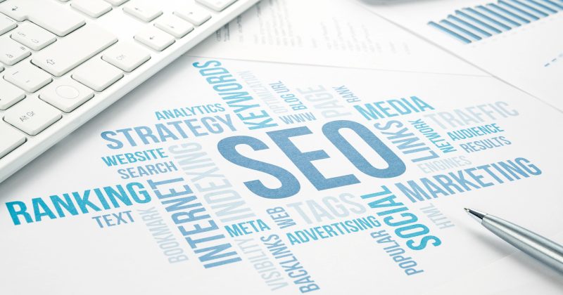 A printed paper with the word "SEO" in large letters, surrounded by terms like "RANKING," "KEYWORDS," and "MARKETING," sits next to a keyboard, pen, calculator, and hints at tools like plugins or WordPress for enhanced optimization.
