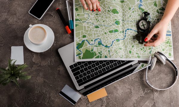 A person’s hands hold a compass and unfold a map over a laptop on a desk with a coffee cup, smartphone, pens, headphones, cards, and a small plant, exploring Interactive Maps or Map Plugins for WordPress.