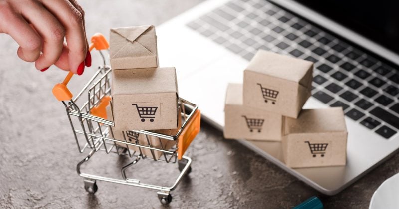 A hand places a small box into a miniature shopping cart near a laptop, with more small boxes featuring shopping cart icons in the background.