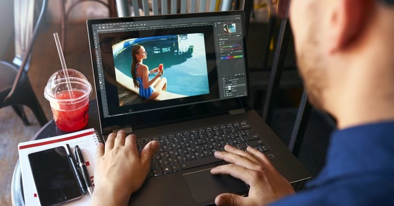 Person editing a photo of a woman by a pool on a laptop, using an Image Optimization Plugin to speed up your site, with a smartphone, notebook, pen, and a red drink on the table nearby.