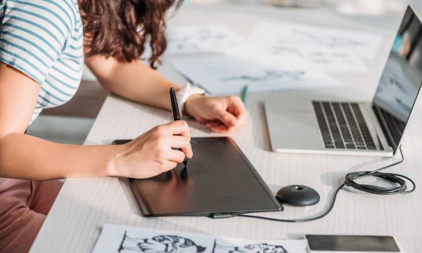 A person using a digital drawing tablet and stylus at a desk with a laptop and character sketches on paper, creating stunning visuals perfect for enhancing WordPress projects or showcasing artwork in Lightbox plugins.