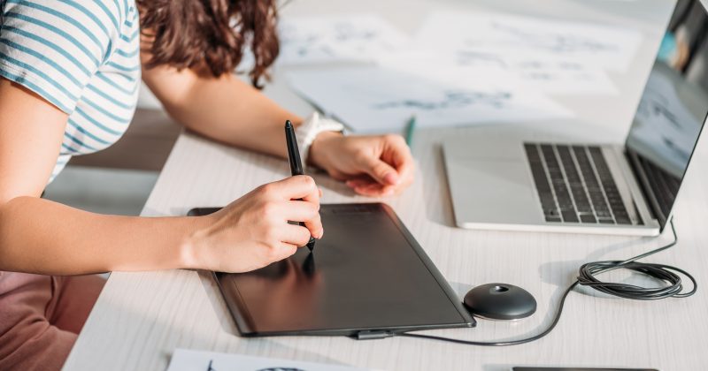 A person using a digital drawing tablet and stylus at a desk with a laptop and character sketches on paper, creating stunning visuals perfect for enhancing WordPress projects or showcasing artwork in Lightbox plugins.