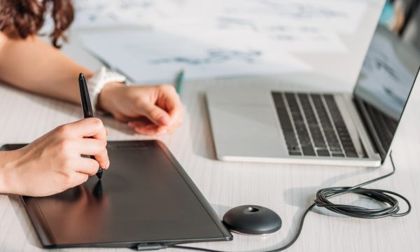 A person using a digital drawing tablet and stylus at a desk with a laptop and character sketches on paper, creating stunning visuals perfect for enhancing WordPress projects or showcasing artwork in Lightbox plugins.