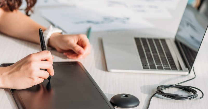 A person using a digital drawing tablet and stylus at a desk with a laptop and character sketches on paper, creating stunning visuals perfect for enhancing WordPress projects or showcasing artwork in Lightbox plugins.