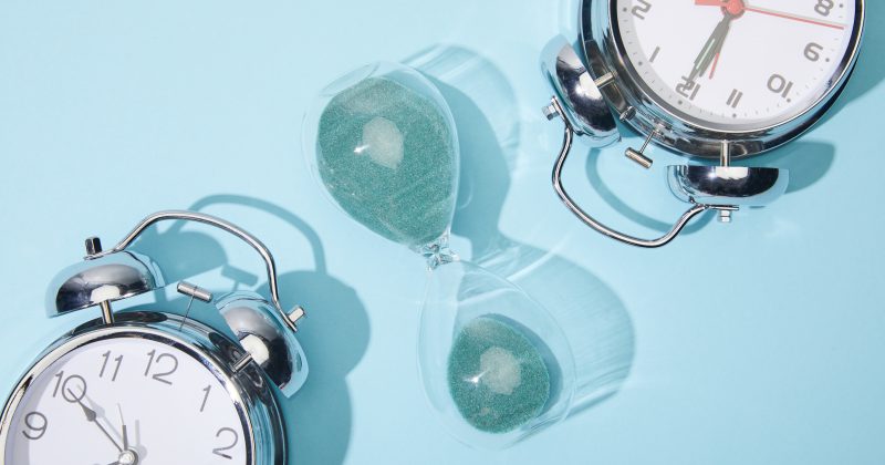 Two silver alarm clocks and a glass hourglass with green sand are placed on a light blue background.