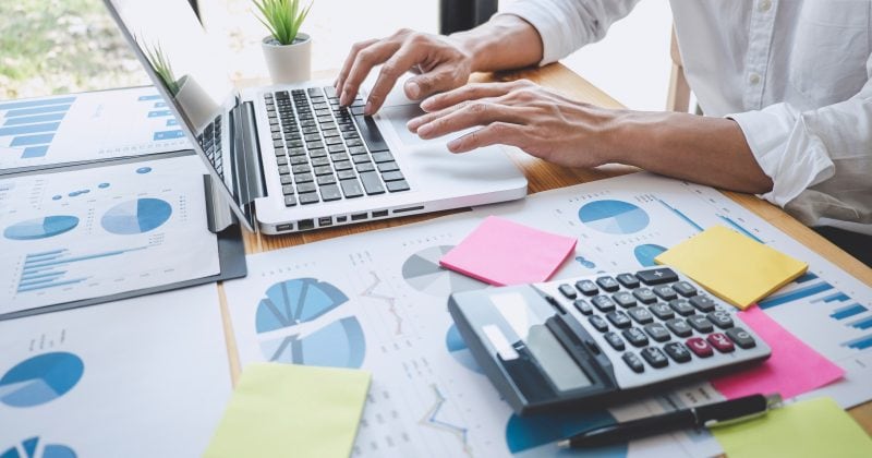 A person works on a laptop at a desk covered with financial charts, a calculator, a pen, and colorful sticky notes.