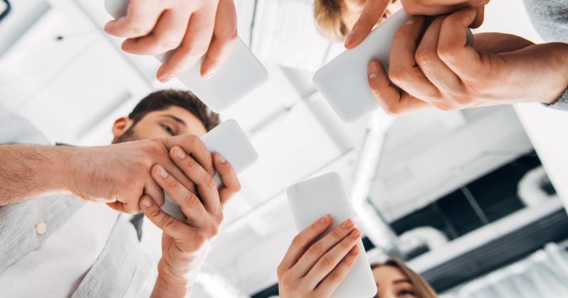Low-angle view of several people standing in a circle, each using a smartphone—perhaps exploring Instagram plugins or ways to display feed on WordPress—with a modern ceiling in the background.