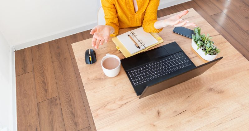 Person in a yellow shirt sits at a wooden desk with a laptop, notebook, coffee mug, mouse, phone, and a small potted plant, gesturing with open hands.