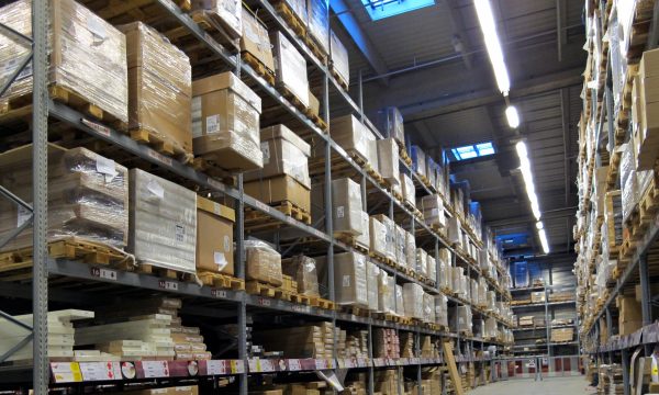 Tall shelves in a warehouse filled with stacked pallets and boxes, with clear aisles and overhead lighting.