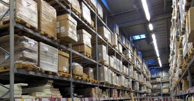 Tall shelves in a warehouse filled with stacked pallets and boxes, with clear aisles and overhead lighting.